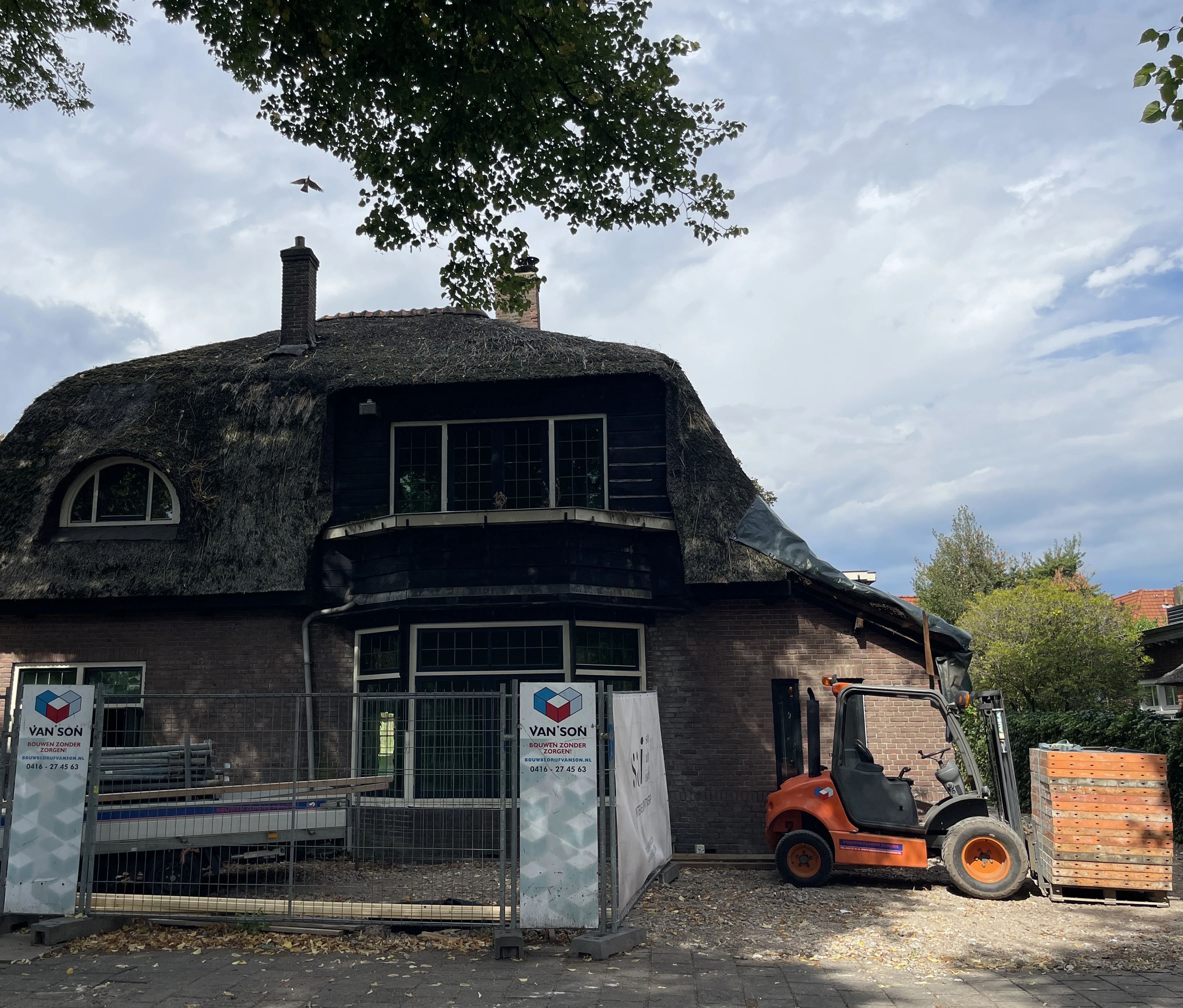 De voorgevel van de villa achter bouwhekken, met een heftruck en pallets op het terrein