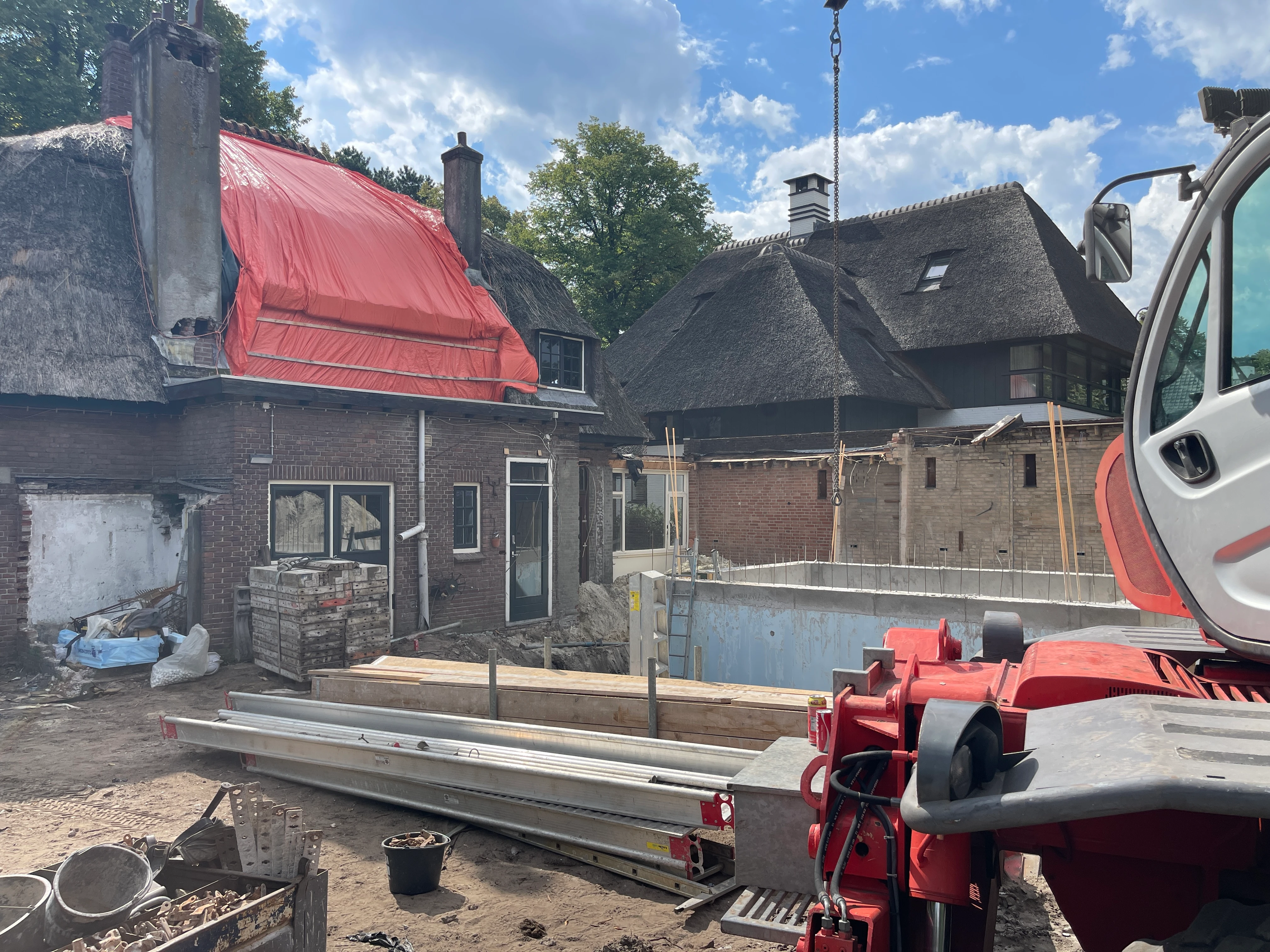 De achterzijde van het huis met een kraan, stalen liggers en de betonnen kelderbak in de bouwput