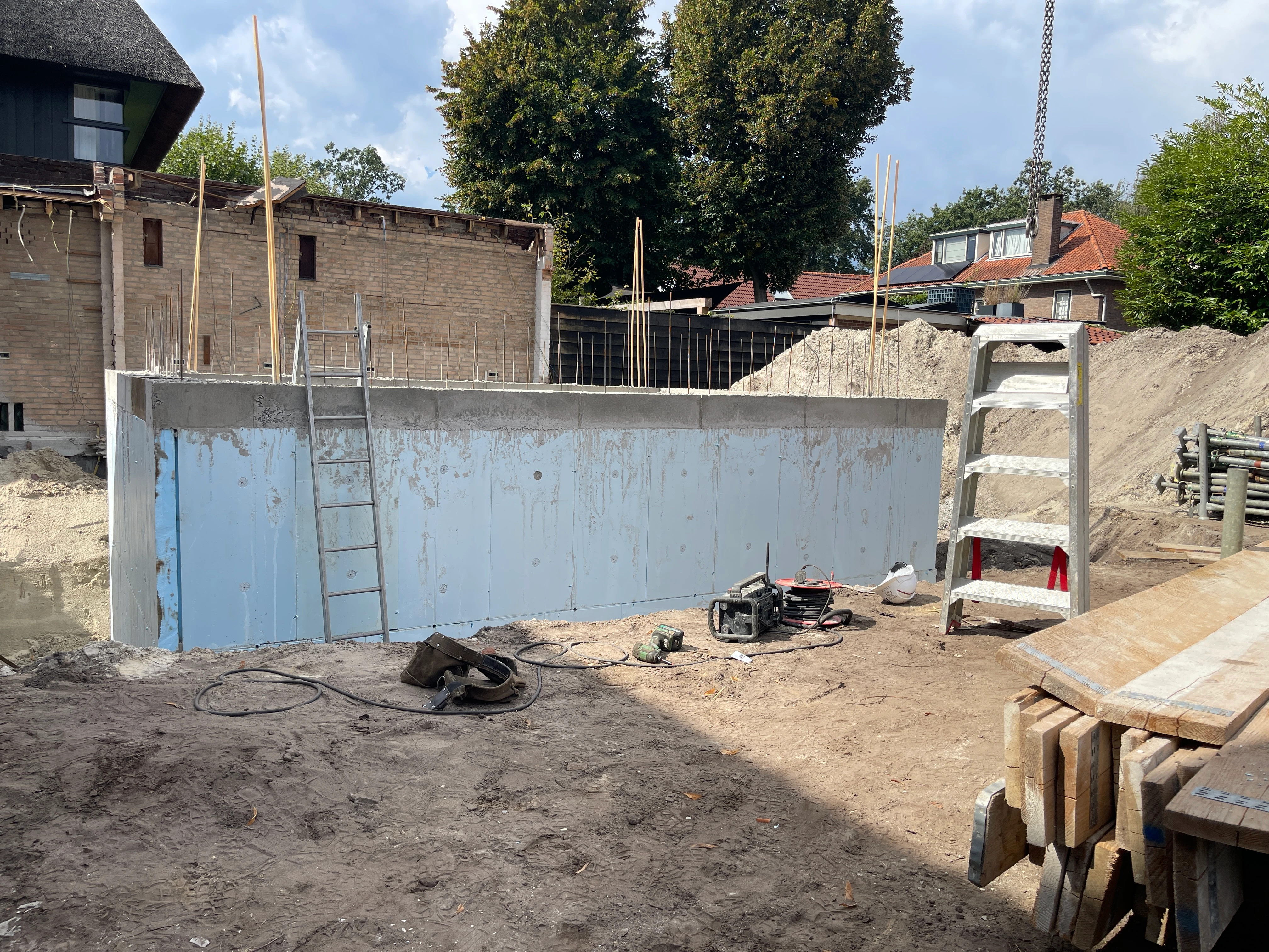 De betonnen kelderwand met wapeningsstaven in de open bouwput naast het bestaande bijgebouw