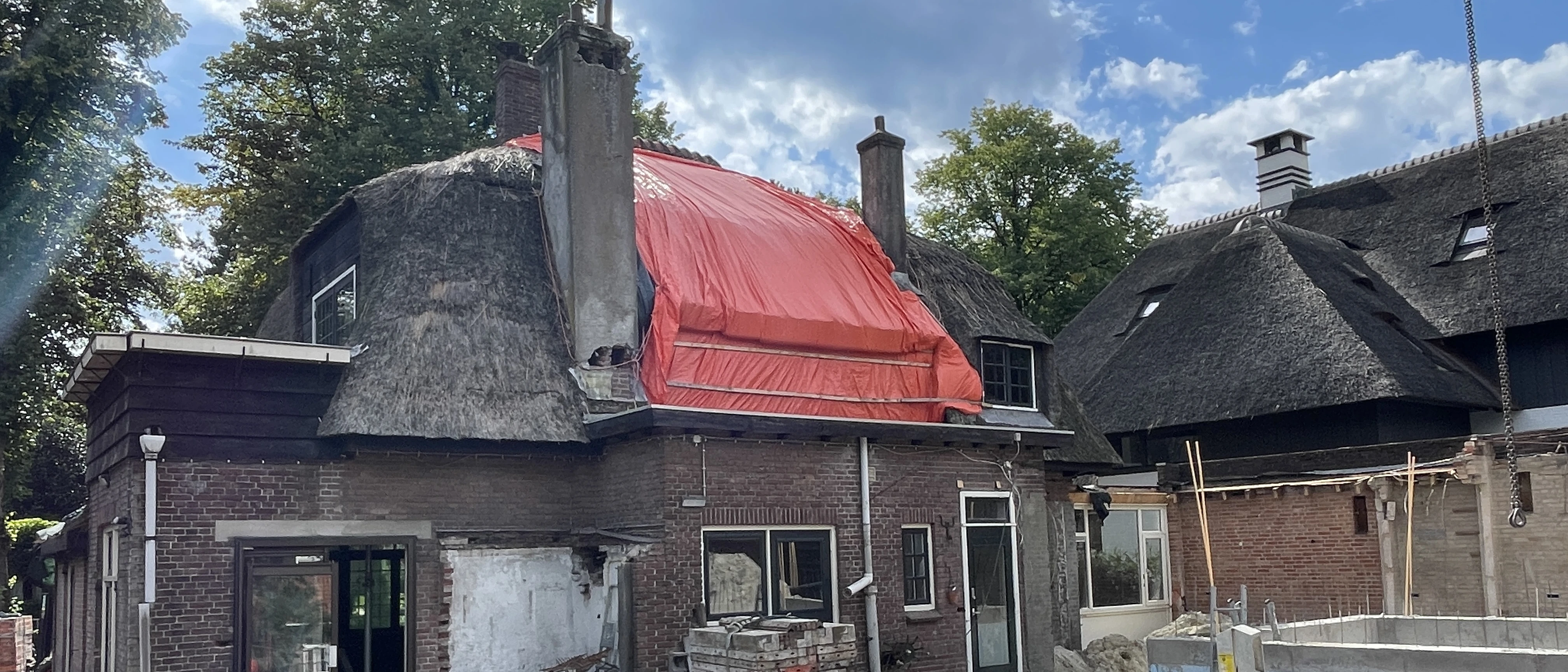 Achterzijde van de rietgedekte villa tijdens de verbouwing, met een rood dekzeil over de kap en gestript metselwerk
